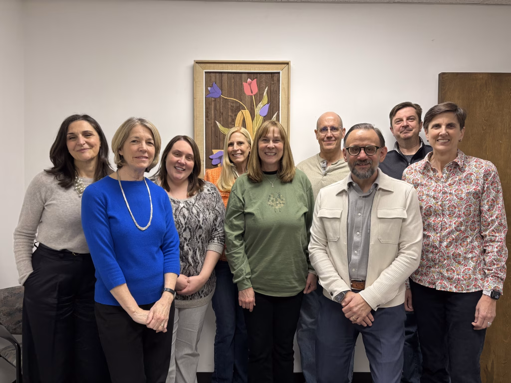 Nine adults stand indoors in front of a white wall and a framed floral artwork, smiling at the camera. Most are wearing casual clothes, and the group appears friendly and relaxed.
