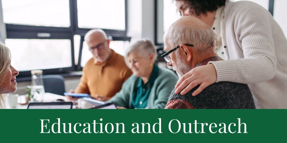A group of older adults sits together at a table, talking and working with papers and tablets. A younger person stands beside them, resting a hand on one man’s shoulder. Text reads “Education and Outreach.”.