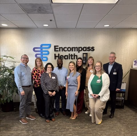 A group of nine professionally dressed people stand and smile in front of an Encompass Health sign inside an office lobby. A potted plant and office furniture are visible in the background.