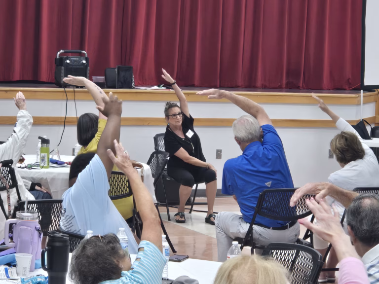 A group of adults seated around tables follow a woman in the center leading a seated stretching exercise, all reaching one arm overhead in a bright room with red curtains.