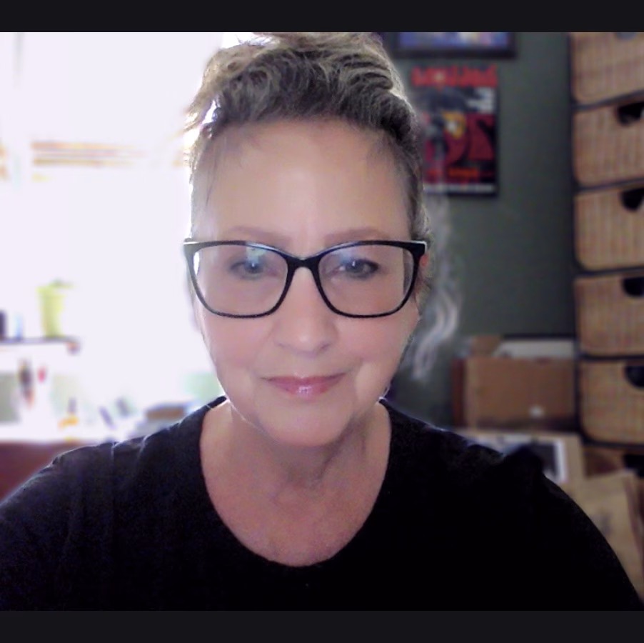 A woman with glasses and light hair pulled up in a bun smiles softly at the camera. She is indoors, with shelves, woven baskets, and posters visible in the softly lit background.