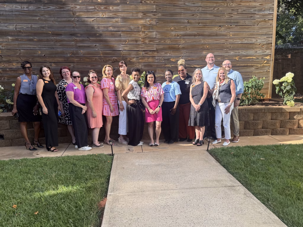 A group of 15 adults pose together outside on a sunny day, standing on a sidewalk in front of a wooden wall and garden. They are smiling and dressed in a mix of casual and business casual clothes.