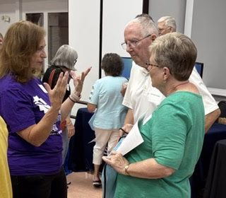 A woman in a purple shirt gestures while talking to an older man and woman who are listening attentively. Several other people are visible in the background of the room.