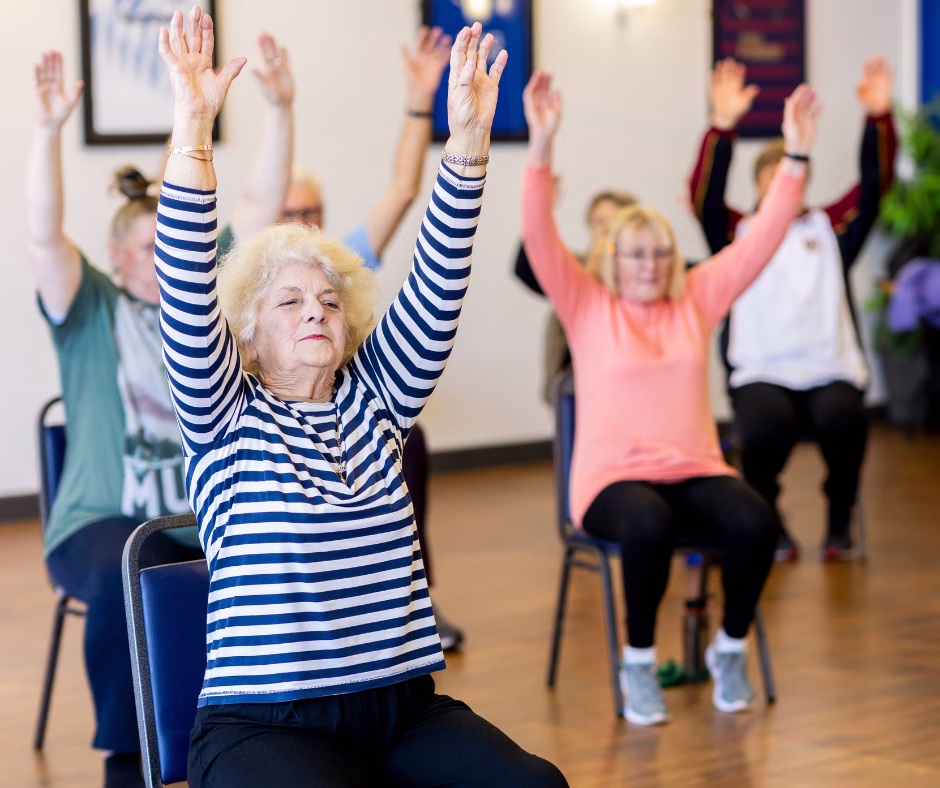 A group of older adults sits on chairs in a bright room, raising their arms overhead as part of a group exercise or fitness class. The participants appear focused and engaged.