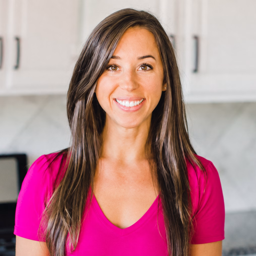 Woman with long brown hair wearing a bright pink top, smiling and standing in a modern kitchen with white cabinets and a tiled backsplash.