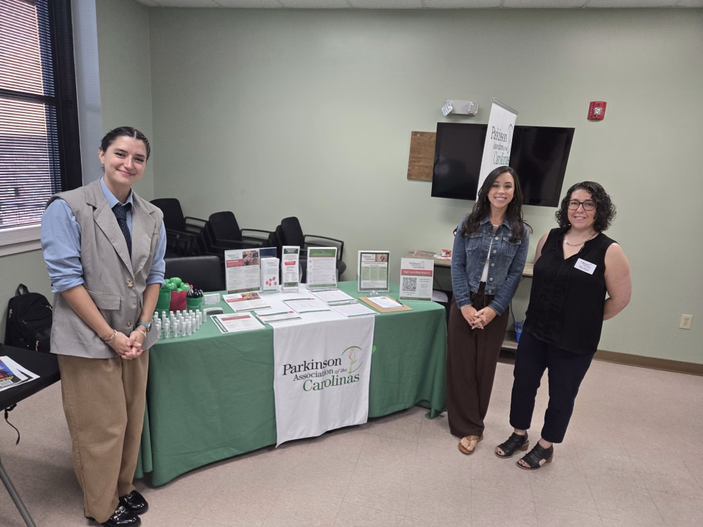 Three women stand and smile in front of an informational table with brochures and a green Parkinson Association of the Carolinas tablecloth in a well-lit room.