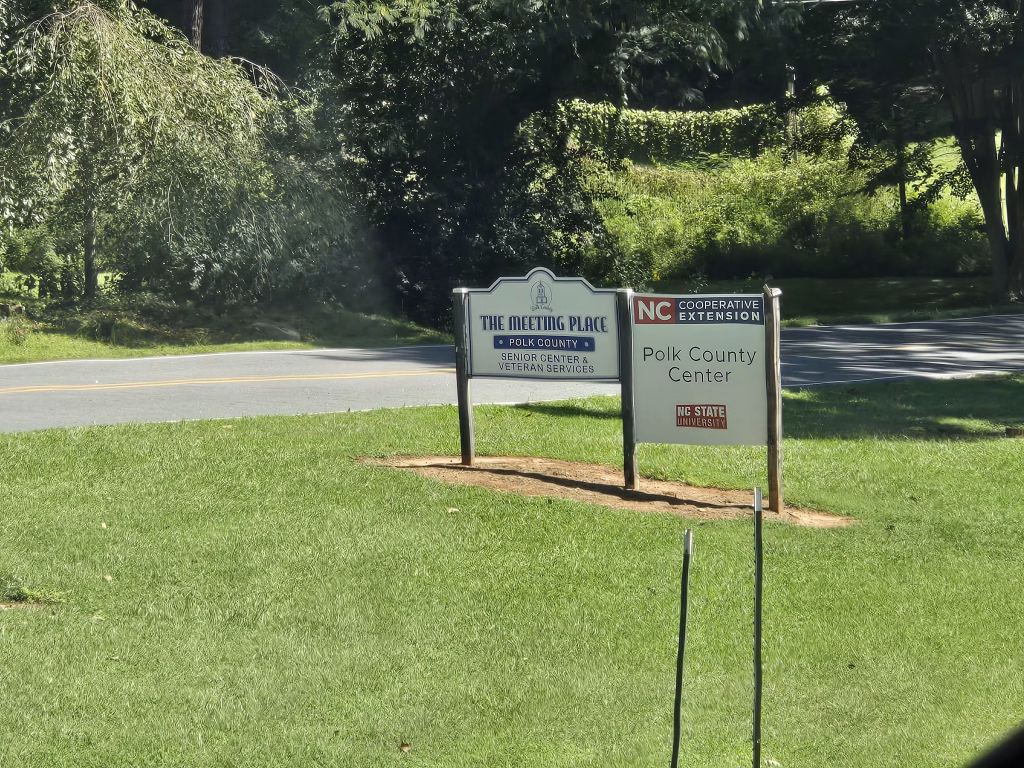 Two signs on a grassy area by a road: one reads The Meeting Place Polk County Senior Center & Veteran Services, the other reads NC Cooperative Extension Polk County Center NC State University. Trees are in the background.