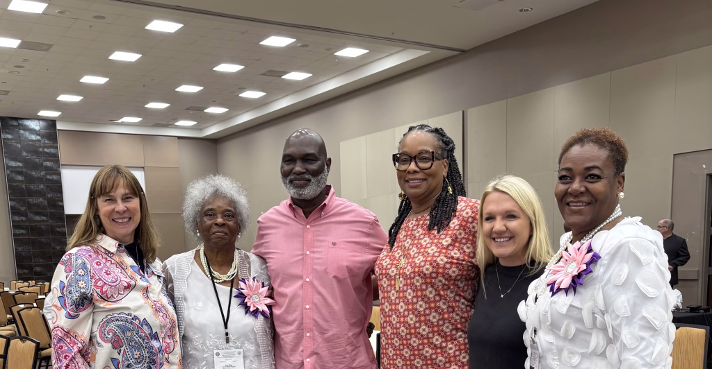 Six people stand side by side, smiling in a large conference room with rows of chairs and beige walls, dressed in colorful and patterned clothing.