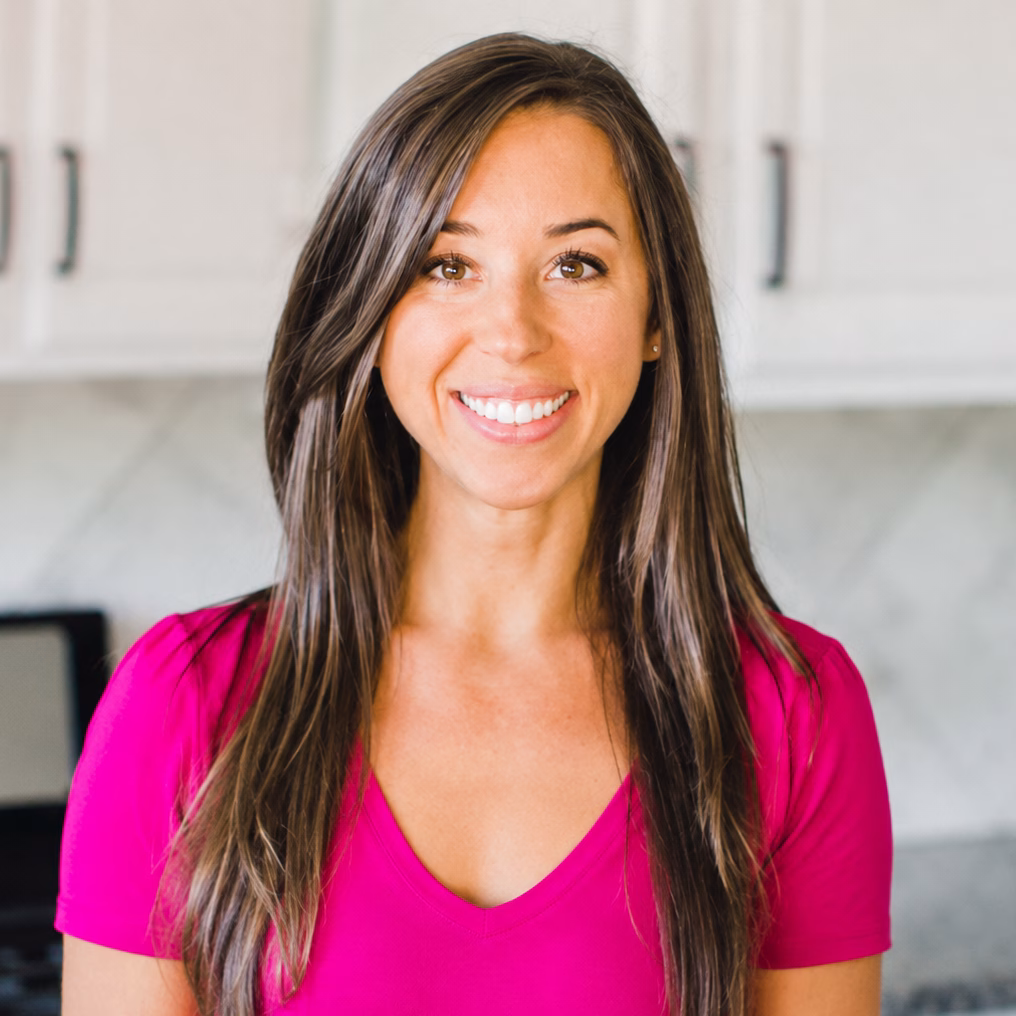 A woman with long brown hair smiles at the camera, wearing a bright pink top. She stands in a kitchen with white cabinets and a tiled backsplash.
